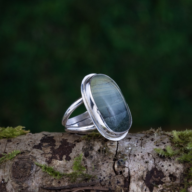 A sterling silver ring set with a hand-cut banded chert cabochon in soft greens and greys, on a mossy log.