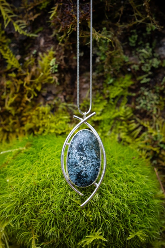 A blue-black brecciated dallasite cabochon in a leaf-shaped sterling silver wire frame, hanging over bright moss.