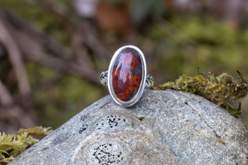 A sterling silver ring with a hand-cut orange peel agate cabochon — deep red plume patterns in the stone — resting on a lichen-covered rock.